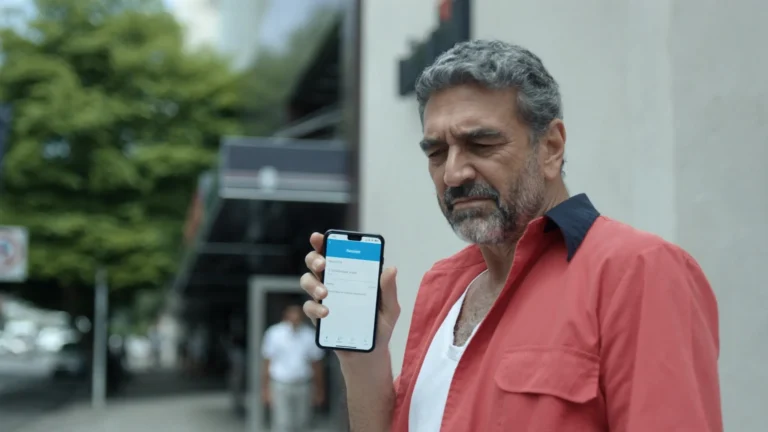 Homem de camisa vermelha na calçada, em frente a um prédio comercial, olha preocupado a tela do celular