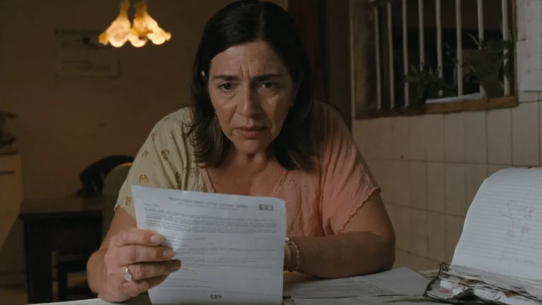 Início Mulher à mesa da cozinha lendo a decisão que limitou os descontos, com o contracheque e anotações de contas ao lado